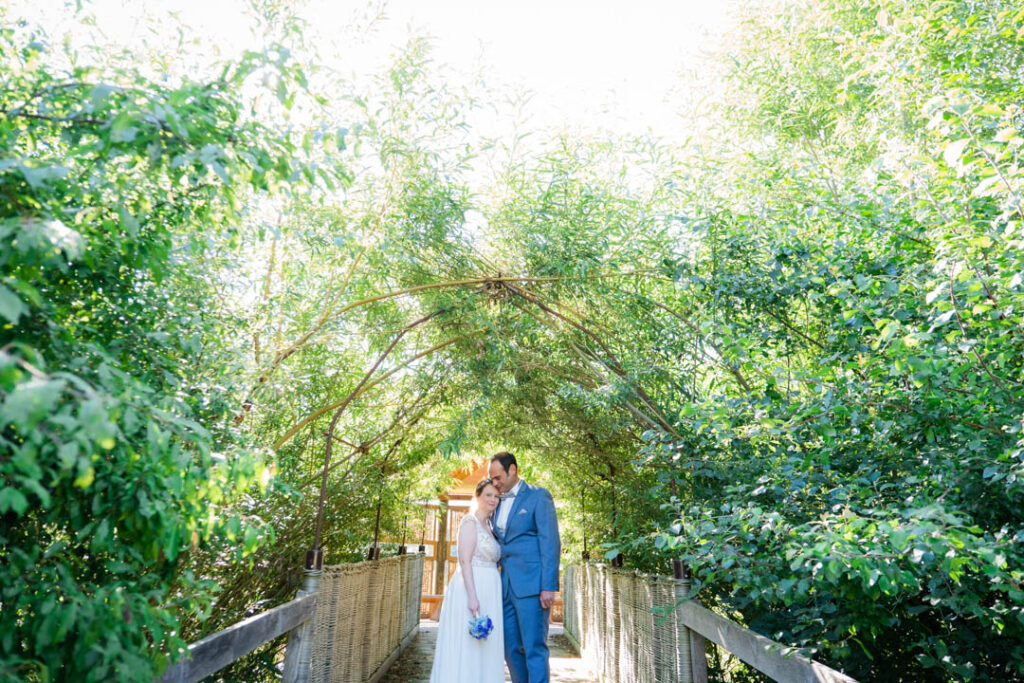 Brautpaar steht unter grünem Blättertunnel auf einer Brücke und umarmt sich